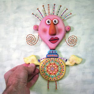May include: A whimsical, handmade figurine with a pink heart-shaped head, large eyes, a red mouth, and a long orange nose. The figurine has yellow wings, a colourful patterned torso, and is standing on small legs.