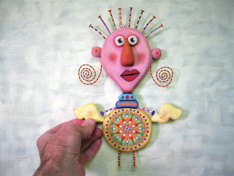 May include: A whimsical, handmade figurine with a pink heart-shaped head, large eyes, a red mouth, and a long orange nose. The figurine has yellow wings, a colourful patterned torso, and is standing on small legs.