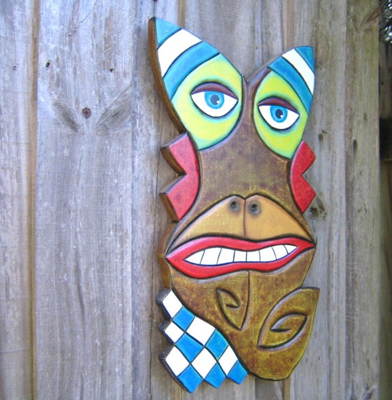 May include: A whimsical, hand-painted wooden mask with a brown base color. The mask features blue eyes, a wide red smiling mouth, and accents of green, red, and blue. A blue and white checkered pattern is on the lower left. The mask is mounted on a wooden surface.