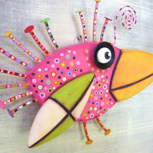May include: A whimsical pink bird sculpture with a long orange beak, green and white wings, and a large black eye. The bird is decorated with colorful polka dots and has a long, curly tail.