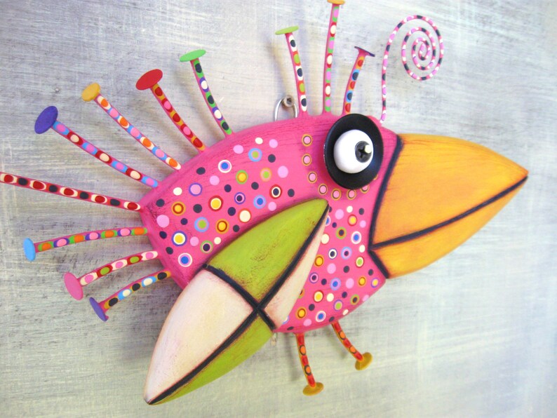 May include: A whimsical pink bird sculpture with a long orange beak, green and white wings, and a large black eye. The bird is decorated with colorful polka dots and has a long, curly tail.