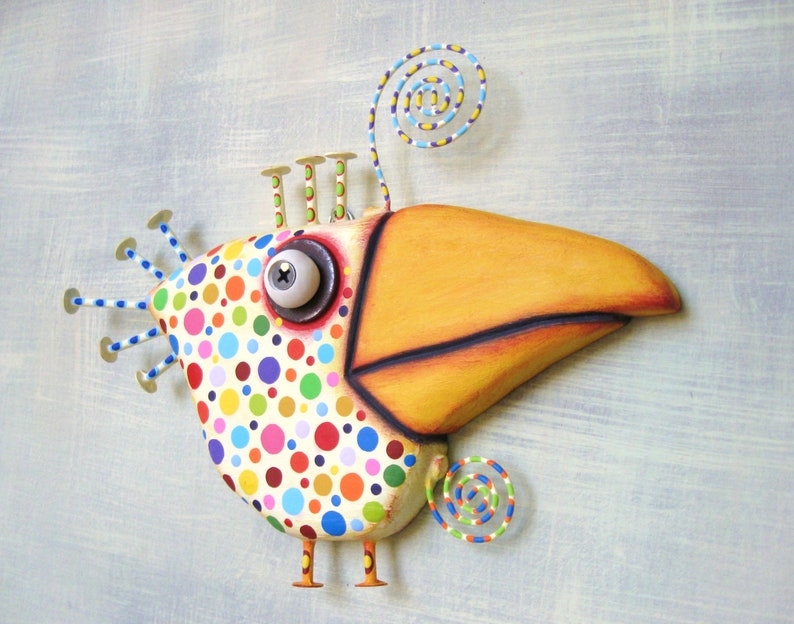 May include: A whimsical, colorful bird sculpture with a large orange beak and a body covered in polka dots. The bird has a large, expressive eye and decorative wire accents. The sculpture is mounted on a light blue background.