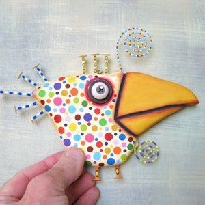 May include: A whimsical, handcrafted bird sculpture with a large yellow beak and a body covered in colorful polka dots. The bird has a large, expressive eye and decorative wire accents. The sculpture is held by a hand against a light blue background.