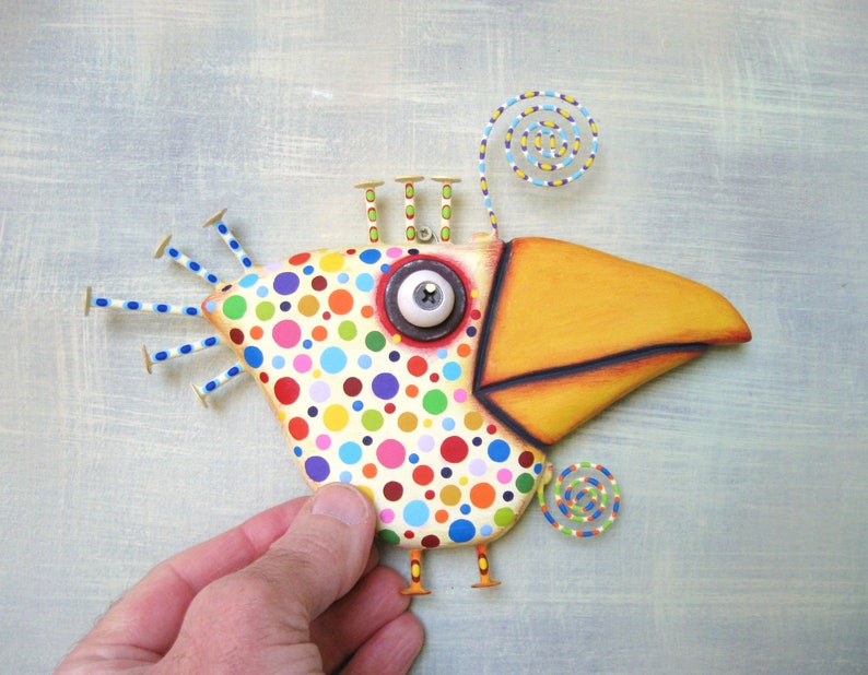 May include: A whimsical, handcrafted bird sculpture with a large yellow beak and a body covered in colorful polka dots. The bird has a large, expressive eye and decorative wire accents. The sculpture is held by a hand against a light blue background.