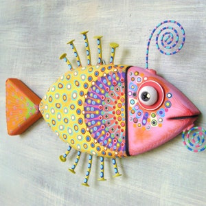 Puede incluir: Una escultura decorativa de pez con un dise&ntilde;o brillante y juguet&oacute;n. El pez tiene una cabeza rosa, un cuerpo amarillo con c&iacute;rculos azules, rojos y morados, y una cola naranja. Cuenta con detalles met&aacute;licos y un ojo grande.