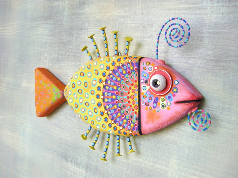 Puede incluir: Una escultura decorativa de pez con un dise&ntilde;o brillante y juguet&oacute;n. El pez tiene una cabeza rosa, un cuerpo amarillo con c&iacute;rculos azules, rojos y morados, y una cola naranja. Cuenta con detalles met&aacute;licos y un ojo grande.