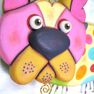 Puede incluir: Una colorida decoraci&oacute;n de pared con forma de perro. La cara del perro est&aacute; pintada en rosa, amarillo y negro, con ojos grandes y expresivos. El cuerpo presenta un patr&oacute;n de lunares multicolores. Decorativo y juguet&oacute;n.