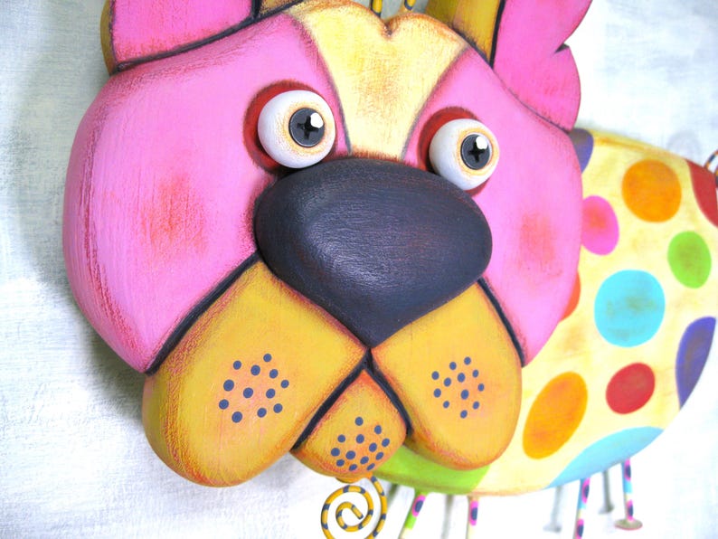 Puede incluir: Una colorida decoraci&oacute;n de pared con forma de perro. La cara del perro est&aacute; pintada en rosa, amarillo y negro, con ojos grandes y expresivos. El cuerpo presenta un patr&oacute;n de lunares multicolores. Decorativo y juguet&oacute;n.