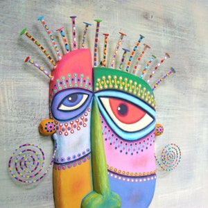 May include: A colorful, abstract mask with a green, pink, and blue face. The mask has two large eyes with white pupils and black outlines. The mask is decorated with small, colorful dots and swirls. The mask is made of wood and has a textured surface.