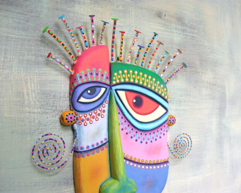 May include: A colorful, abstract mask with a green, pink, and blue face. The mask has two large eyes with white pupils and black outlines. The mask is decorated with small, colorful dots and swirls. The mask is made of wood and has a textured surface.