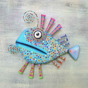 May include: A whimsical, hand-painted fish sculpture in shades of turquoise, adorned with colorful polka dots and spiral accents. The fish features a large eye, fins, and legs, creating a playful, artistic design. This decorative piece is perfect for adding a touch of whimsy to any space.