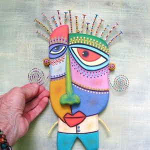 May include: A colorful, abstract face sculpture with a green nose, red lips, and blue eyes. The sculpture is made of painted wood and has a textured surface. It is decorated with colorful beads and spirals.
