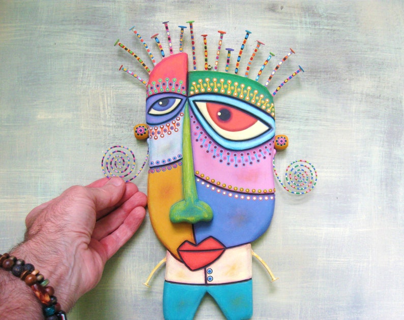 May include: A colorful, abstract face sculpture with a green nose, red lips, and blue eyes. The sculpture is made of painted wood and has a textured surface. It is decorated with colorful beads and spirals.