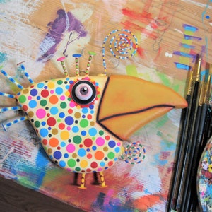 May include: A whimsical, colorful bird sculpture with a large yellow beak and a body covered in polka dots. The bird has a large eye and decorative accents. Paintbrushes and a palette are visible in the background, suggesting an art theme.