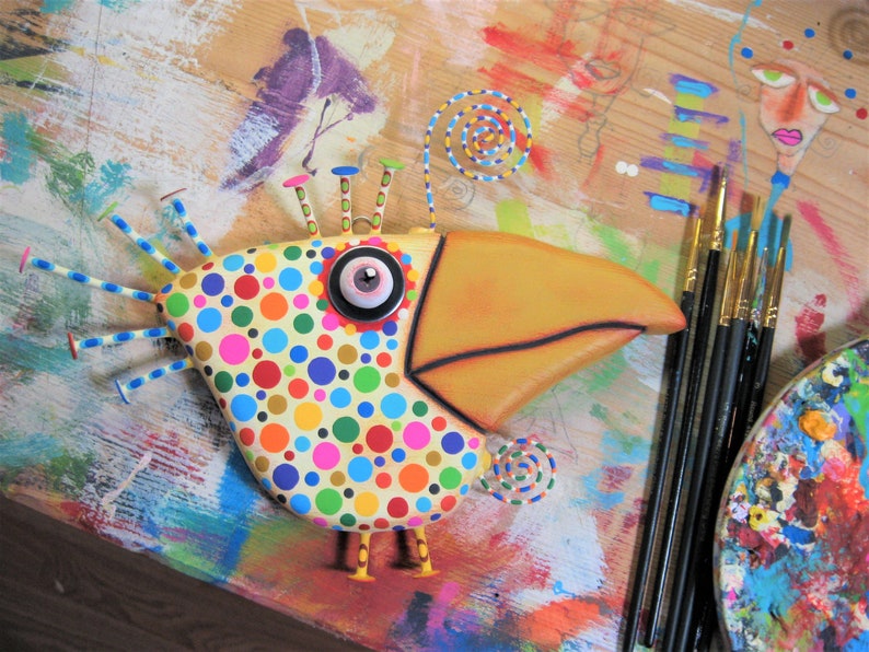 May include: A whimsical, colorful bird sculpture with a large yellow beak and a body covered in polka dots. The bird has a large eye and decorative accents. Paintbrushes and a palette are visible in the background, suggesting an art theme.