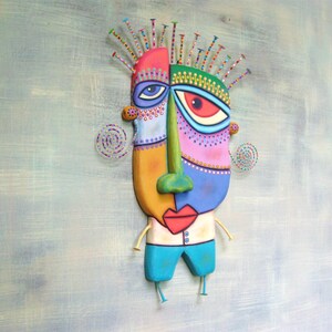 May include: A colorful, abstract face sculpture with a whimsical design. The face is made of painted wood and has a green nose, blue eyes, and a red mouth. The sculpture is attached to a wooden base with legs.