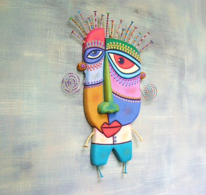 May include: A colorful, abstract face sculpture with a whimsical design. The face is made of painted wood and has a green nose, blue eyes, and a red mouth. The sculpture is attached to a wooden base with legs.
