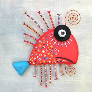 May include: A red, whimsical fish sculpture with a large black eye, a blue tail fin, and colorful dots. The fish has a spiky, colorful, metal fringe around its body and a spiral tail.
