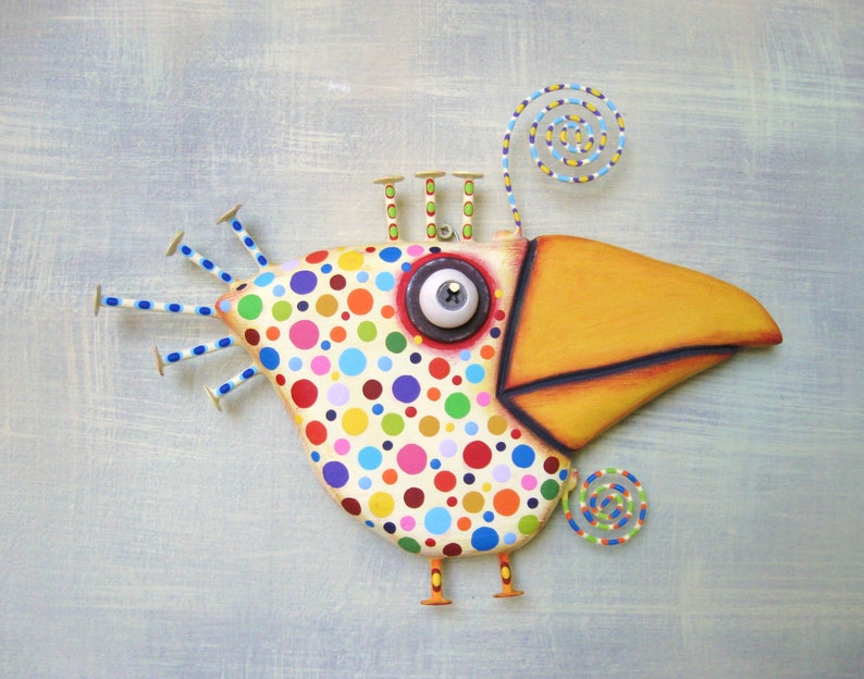 May include: A whimsical, colorful bird sculpture with a large yellow beak and a body covered in polka dots. The bird has a large, expressive eye and nail-like accents. The sculpture is a unique piece of art.