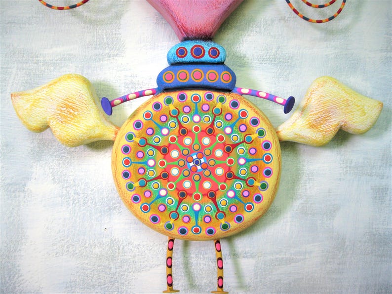 May include: A whimsical wooden sculpture of a bird with yellow wings, a pink head, and a colourful circular body with a mandala design. The bird has blue and pink legs and is painted with a variety of colours, including yellow, pink, blue, green, and red.
