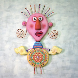 May include: A whimsical, pink and yellow figurine with a large, round head, a long nose, and a colourful, circular body. The figurine has a pair of wings and a crown of colourful spikes on its head.