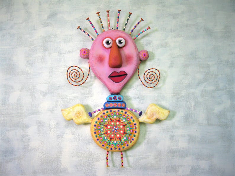 May include: A whimsical, pink and yellow figurine with a large, round head, a long nose, and a colourful, circular body. The figurine has a pair of wings and a crown of colourful spikes on its head.
