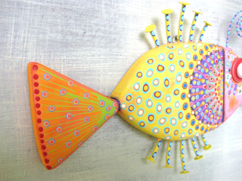 Puede incluir: Una escultura de pez pintada a mano con una paleta de colores vibrantes. El pez presenta un cuerpo amarillo adornado con c&iacute;rculos azules y naranjas, una cola naranja con detalles verdes y azules y una cabeza multicolor. Tiene aletas amarillas y un ojo rojo.