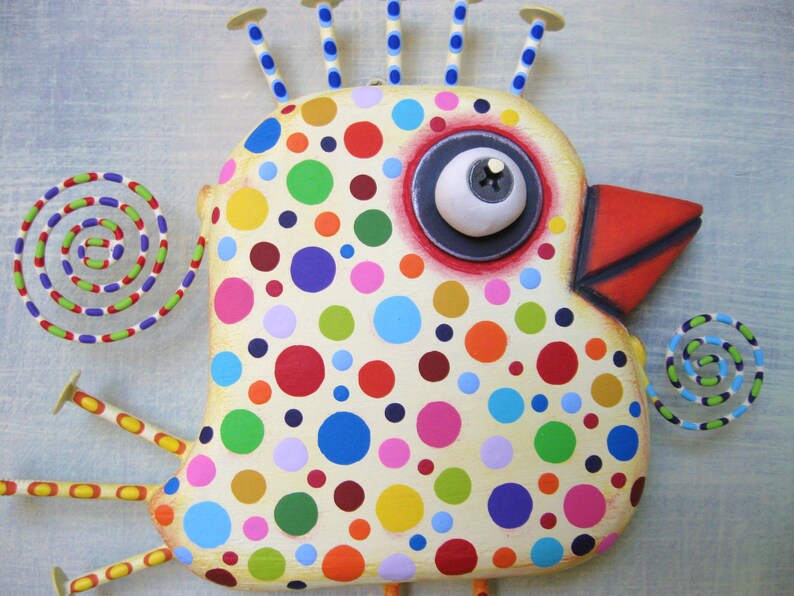 May include: A whimsical, handcrafted bird sculpture with a cream-colored body adorned with colorful polka dots. It features a large eye, a red beak, and decorative spirals. The bird has multiple antennae and legs, all with yellow and blue accents.