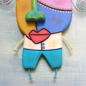 May include: A whimsical wooden sculpture of a face with a yellow, pink, blue, and green color scheme. The face has a large red mouth, a green nose, and a white shirt with two blue buttons. The sculpture has two gold arms and two blue legs.
