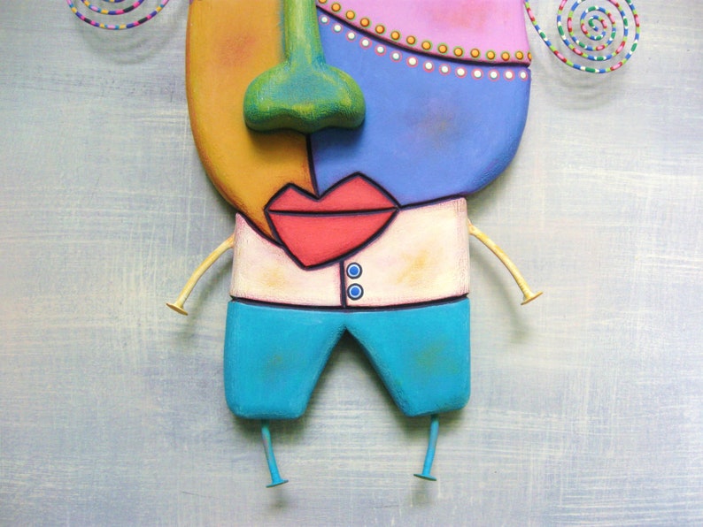 May include: A whimsical wooden sculpture of a face with a yellow, pink, blue, and green color scheme. The face has a large red mouth, a green nose, and a white shirt with two blue buttons. The sculpture has two gold arms and two blue legs.