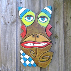 May include: A colorful, wooden tiki mask with a whimsical design. The mask features blue eyes, a wide red mouth, and a brown face with yellow, blue, and white accents. A blue and white checkered pattern is on the lower left side.