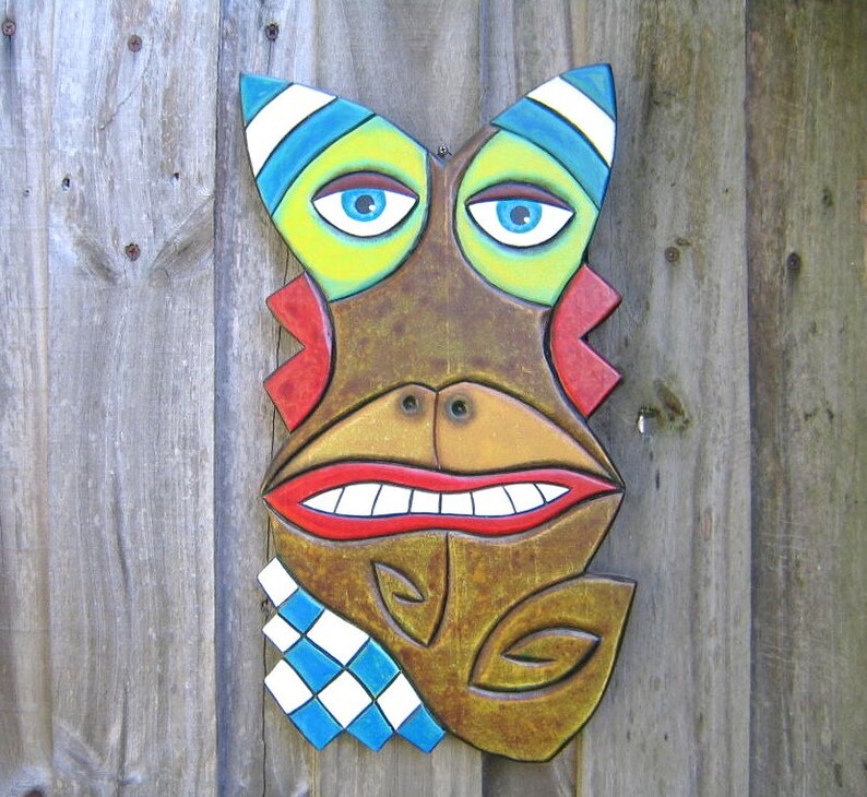 May include: A colorful, wooden tiki mask with a whimsical design. The mask features blue eyes, a wide red mouth, and a brown face with yellow, blue, and white accents. A blue and white checkered pattern is on the lower left side.