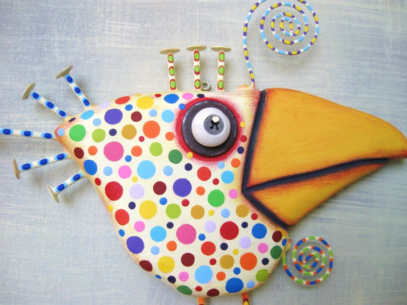 May include: Whimsical, hand-painted bird sculpture with a cream-colored body covered in colorful polka dots. Features a large yellow beak, a single eye, and decorative accents. The bird has a unique, artistic design, perfect for home decor.