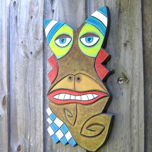 May include: A whimsical, wooden mask with a cartoonish face. The mask features large blue eyes, a wide red mouth, and a brown face with colorful accents of yellow, blue, and red. A blue and white checkered pattern adorns the lower portion.