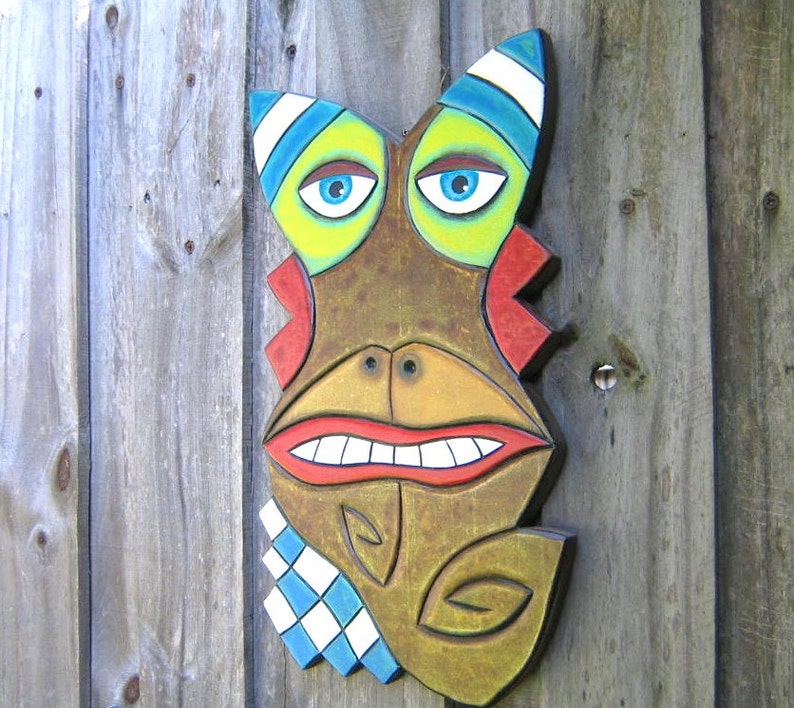 May include: A whimsical, wooden mask with a cartoonish face. The mask features large blue eyes, a wide red mouth, and a brown face with colorful accents of yellow, blue, and red. A blue and white checkered pattern adorns the lower portion.