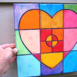 May include: A colorful heart-shaped wall art piece made of wood tiles. The heart is composed of various shades of orange, yellow, pink, red, green, and blue. The tiles are arranged in a geometric pattern, creating a modern and abstract design.