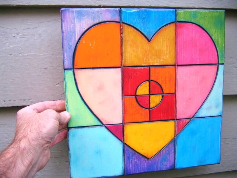 May include: A colorful heart-shaped wall art piece made of wood tiles. The heart is composed of various shades of orange, yellow, pink, red, green, and blue. The tiles are arranged in a geometric pattern, creating a modern and abstract design.