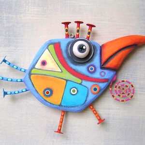 May include: A whimsical, hand-painted wooden bird sculpture. The bird features a blue body with colorful geometric shapes, an orange beak, and a large eye. It has three red antennae and three blue legs with red feet. A decorative spiral is attached.
