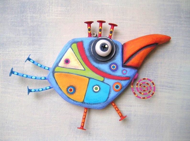 May include: A whimsical, hand-painted wooden bird sculpture. The bird features a blue body with colorful geometric shapes, an orange beak, and a large eye. It has three red antennae and three blue legs with red feet. A decorative spiral is attached.