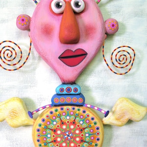 May include: A whimsical, pink and yellow sculpture of a person with large eyes, a long nose, and a red mouth. The sculpture has a colourful, circular design on the bottom and yellow wings.