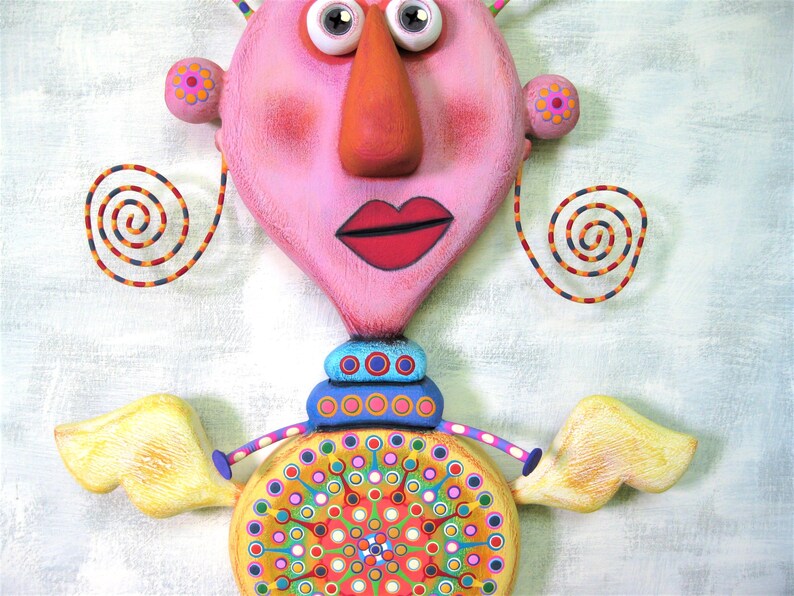 May include: A whimsical, pink and yellow sculpture of a person with large eyes, a long nose, and a red mouth. The sculpture has a colourful, circular design on the bottom and yellow wings.