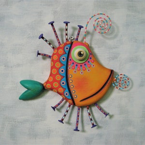 May include: A whimsical, colorful fish sculpture with an orange body, adorned with red and blue patterns. It features a green eye, a green tail, and decorative wire spirals. The fish has purple and white accents, creating a playful, artistic design.