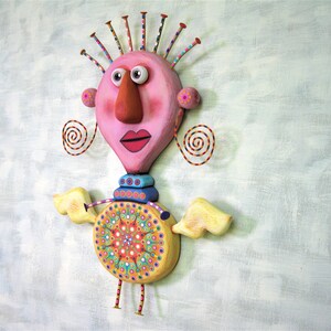 May include: A whimsical, colourful, and textured sculpture of a person with a pink head, a large orange nose, and yellow wings. The sculpture has a circular, multi-coloured design on its torso.