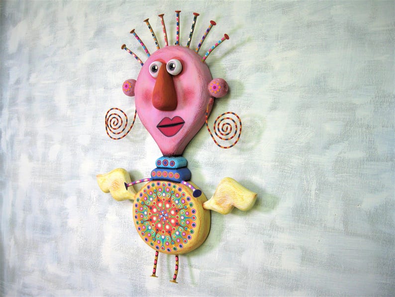 May include: A whimsical, colourful, and textured sculpture of a person with a pink head, a large orange nose, and yellow wings. The sculpture has a circular, multi-coloured design on its torso.