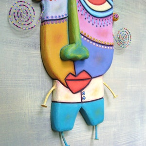 May include: A colorful, whimsical sculpture of a face with a large, green nose, red lips, and blue and pink skin. The sculpture has yellow arms and blue pants and is attached to a light blue background.