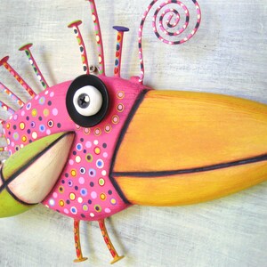 May include: A whimsical, hand-crafted bird sculpture with a long, orange beak, pink body, and a black eye. The bird has a green beak, a white eye, and is decorated with colorful dots and swirls.