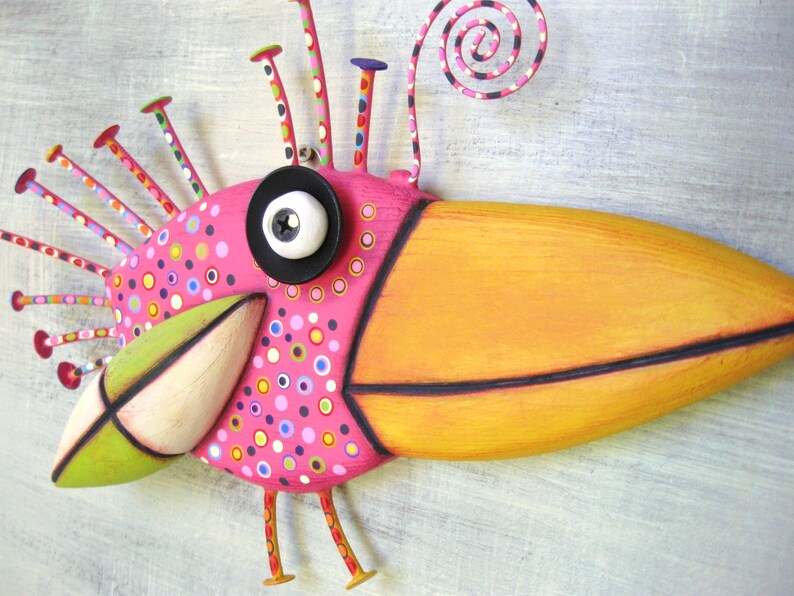 May include: A whimsical, hand-crafted bird sculpture with a long, orange beak, pink body, and a black eye. The bird has a green beak, a white eye, and is decorated with colorful dots and swirls.