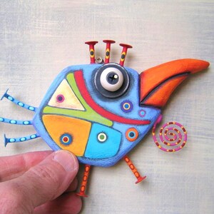 May include: A whimsical, hand-painted bird sculpture with a blue body, orange beak, and red accents. The bird has a large eye, decorative legs, and a spiral tail. The artwork is held by a hand against a light blue background.