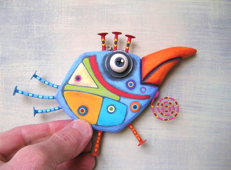 May include: A whimsical, hand-painted bird sculpture with a blue body, orange beak, and red accents. The bird has a large eye, decorative legs, and a spiral tail. The artwork is held by a hand against a light blue background.