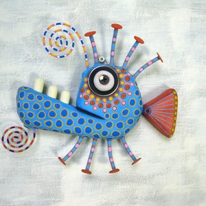 May include: A whimsical blue and yellow fish sculpture with a large eye, a spiral tail, and multiple legs. The fish is decorated with colorful polka dots and has a red fin.